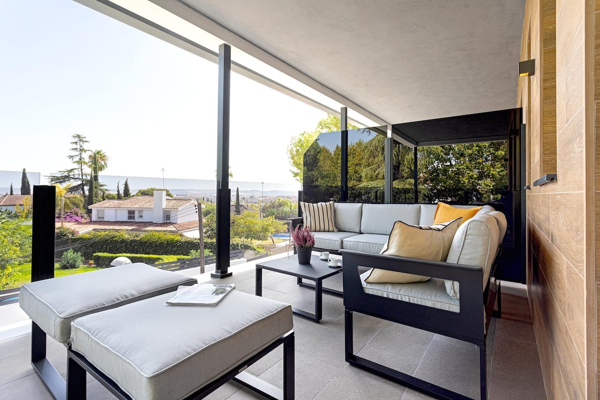 Un espacio exterior moderno con sofá, dos sillones, una mesa baja y vistas a la ciudad y al verdor.