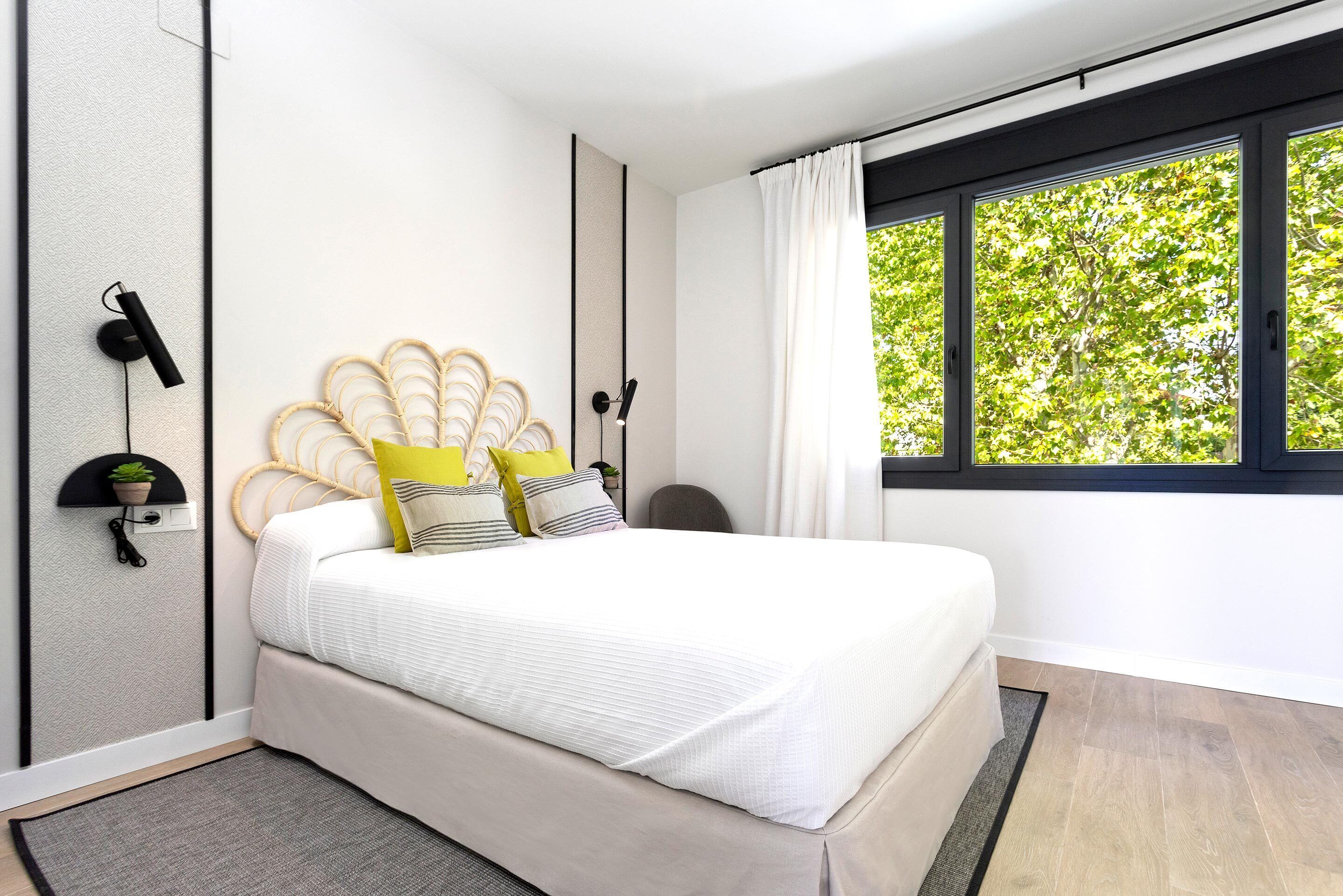 A modern bedroom with a bed, a window with curtains, a wall-mounted lamp, and a small plant.