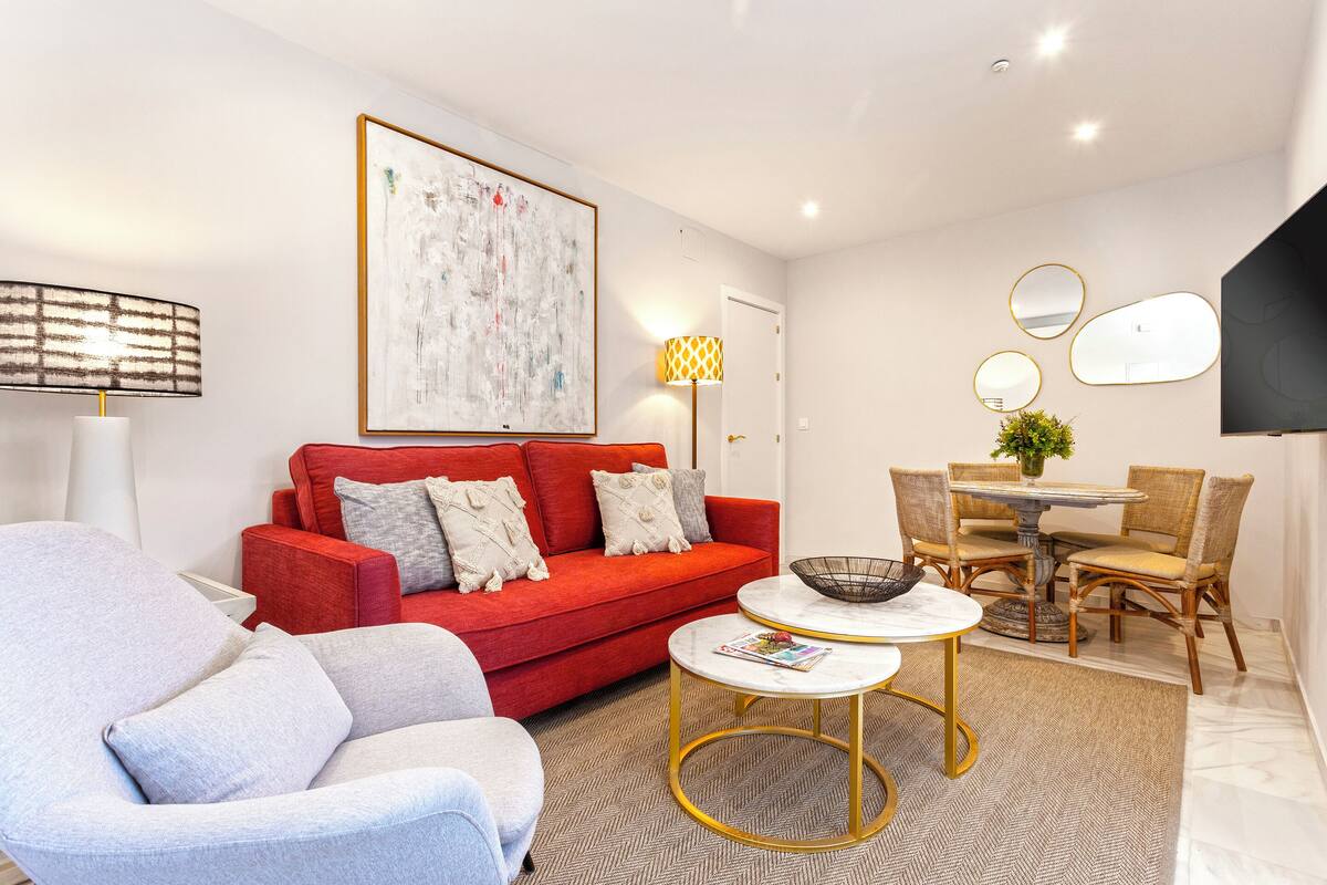 Een moderne woonkamer met een rode bank, een grijze fauteuil, een witte salontafel en een eethoek met rieten stoelen en een tafel.
