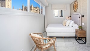 Ein Schlafzimmer mit einem großen Bett, einem Korbstuhl, einem Nachttisch und Blick auf ein historisches Gebäude durch ein Fenster.