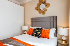 Uma cama bem arrumada com roupa de cama branca e laranja, um encosto de cabeceira estofado em cinza e um espelho decorativo na parede.