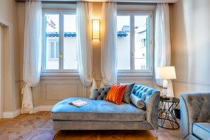 Um quarto com um sofá azul, uma pequena mesa com uma luminária e vista para edifícios através de grandes janelas.