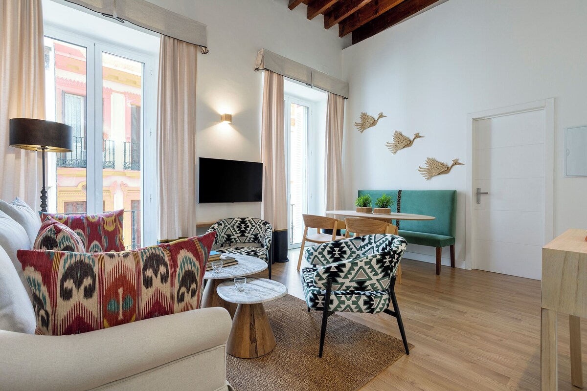 A modern living room with a sofa, chairs, a coffee table, and a TV. There is a balcony with a view of buildings.