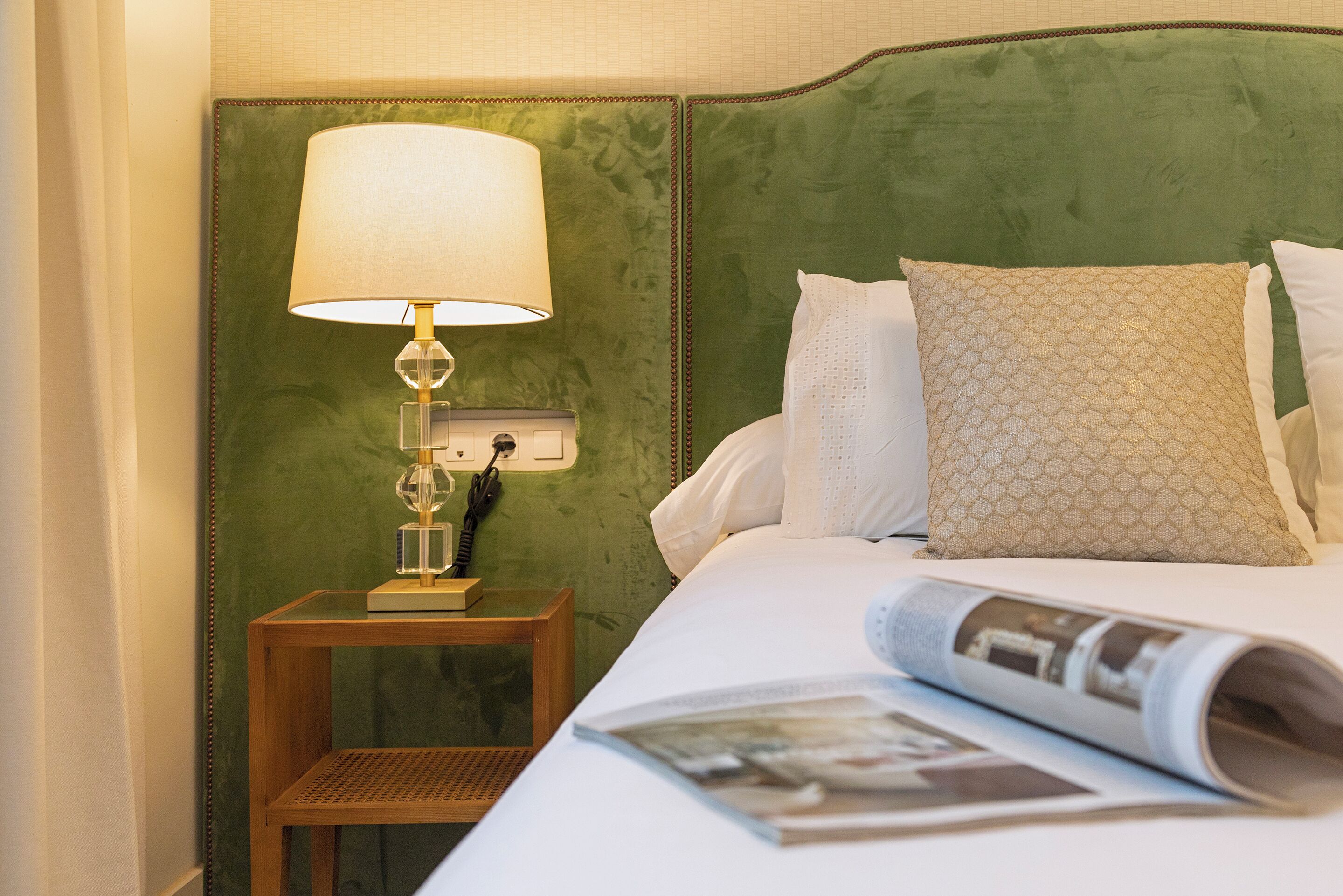 A neatly made bed with a rolled-up magazine, a bedside lamp, and a telephone on a wooden nightstand.