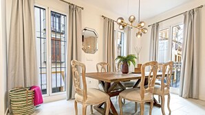 A dining area with a wooden table, six chairs, a chandelier, and large windows with curtains. - Genteel Home Feria (Seville)