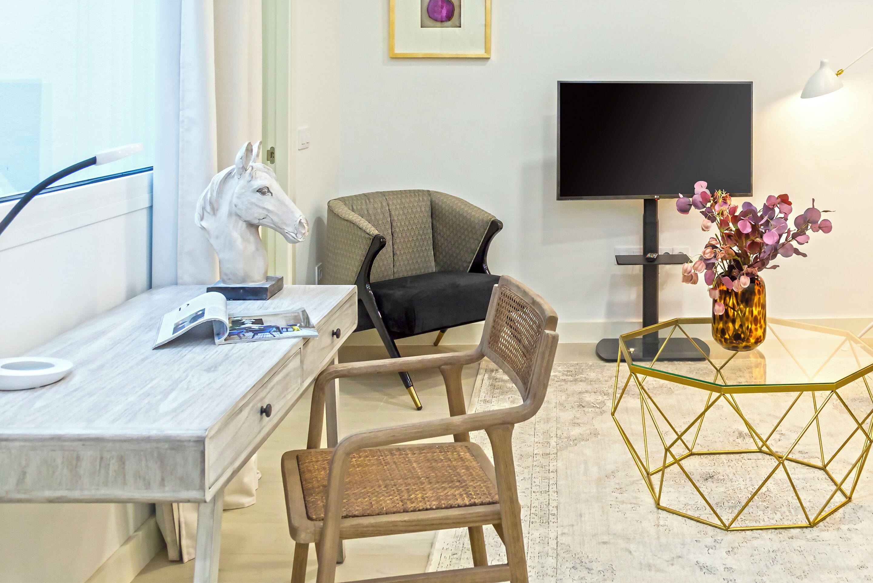 Ett rum med ett skrivbord i trä, en stol, en tv, en vas med blommor och ett geometriskt format soffbord.