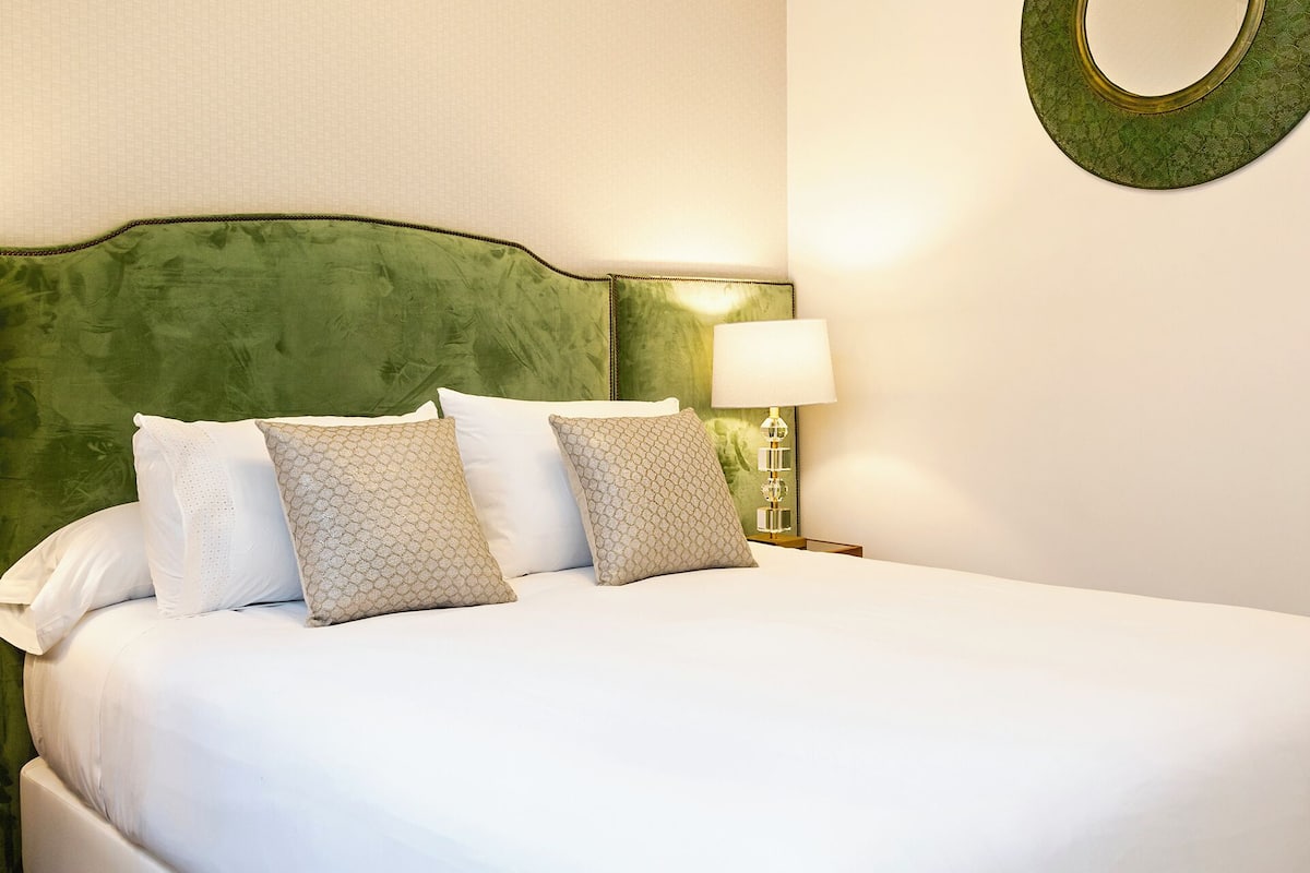 A neatly made bed with white linens and a green upholstered headboard. A round mirror hangs on the wall above the bed.