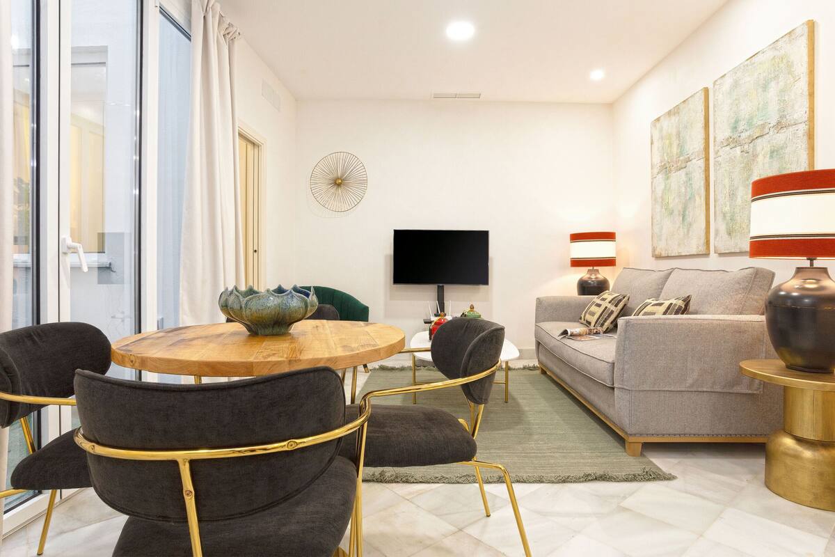 A modern living room with a round wooden table, four upholstered chairs, a grey sofa, a flat-screen TV, and two wall-mounted lamps.