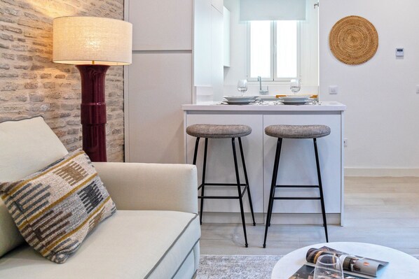 Une cuisine moderne avec un plan de travail blanc, deux tabourets, une lampe suspendue et une décoration murale en fibres tressées.
