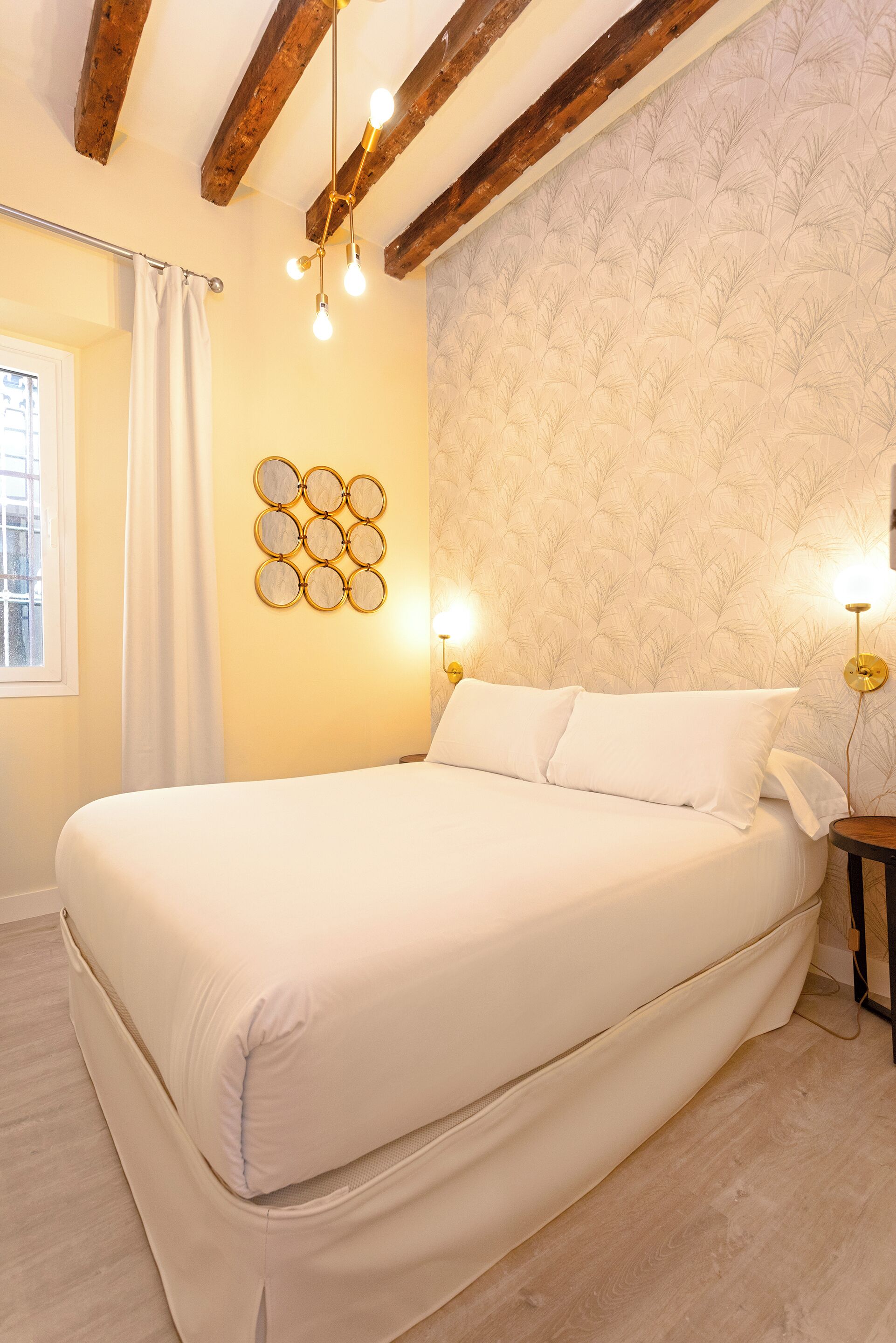 Un dormitorio con una cama grande, vigas de madera, una lámpara de pared y una ventana con cortina.