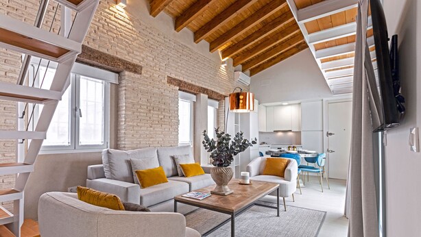 A modern living room with a brick wall, wooden beams, a sofa set, a coffee table, and a dining area in the background. - Genteel Home Feria (Seville)