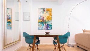 Une pièce moderne avec une table à manger en bois, deux chaises de couleur turquoise et un canapé beige. Des tableaux abstraits ornent le mur, et une lampe sur pied est posée au sol.