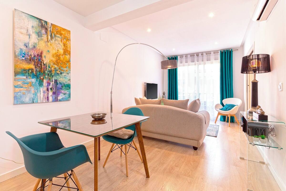 Un salon moderne avec une table à manger en verre, des chaises de couleur turquoise, un canapé beige et un grand tableau abstrait accroché au mur.