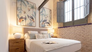 A bedroom with a large bed, two wall-mounted paintings of birds, a nightstand with a lamp, and a brick wall. - Genteel Home Almansa (Seville)