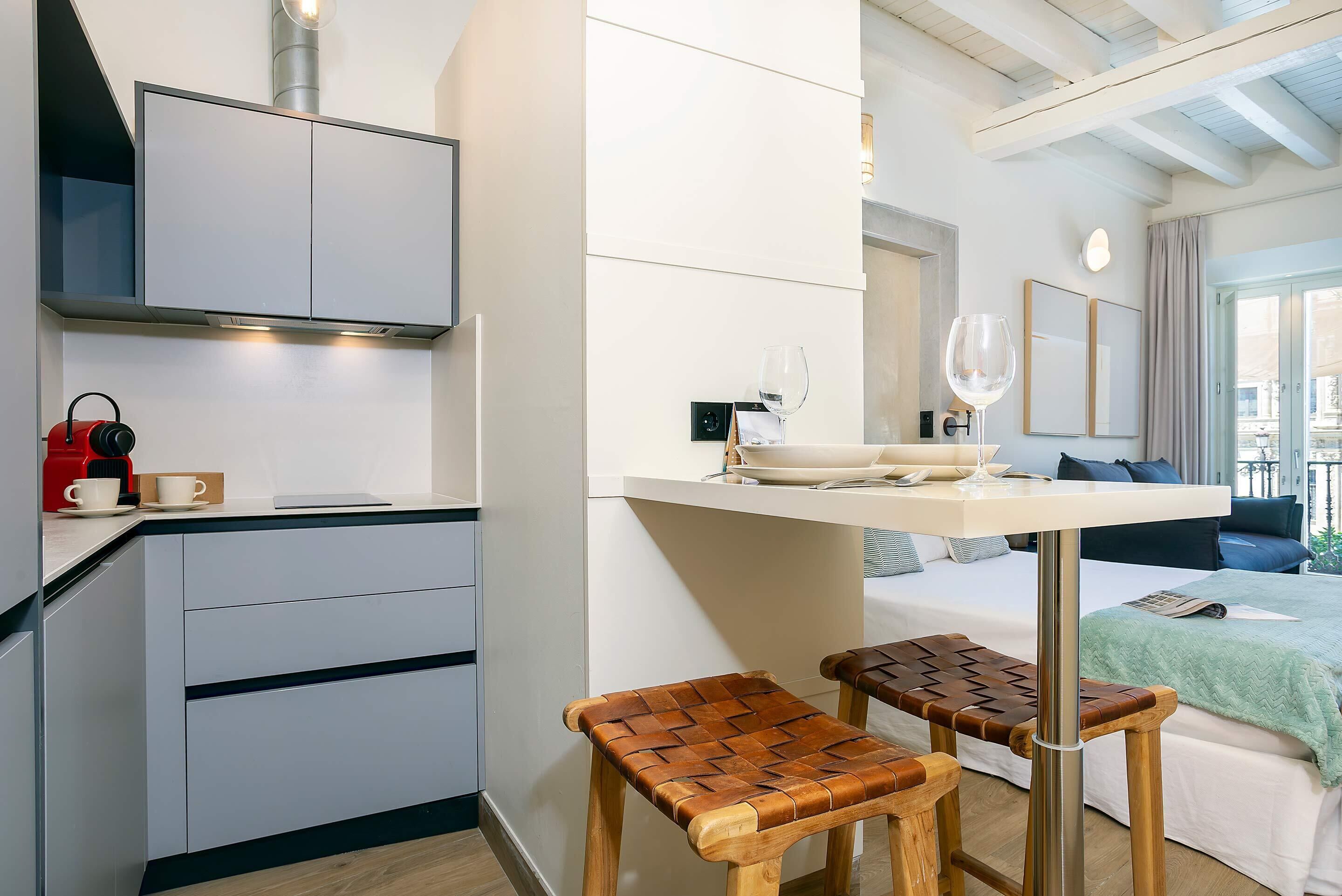 Una cocina moderna con estufa integrada, un comedor con mesa y dos taburetes, y un dormitorio con cama y ventana.