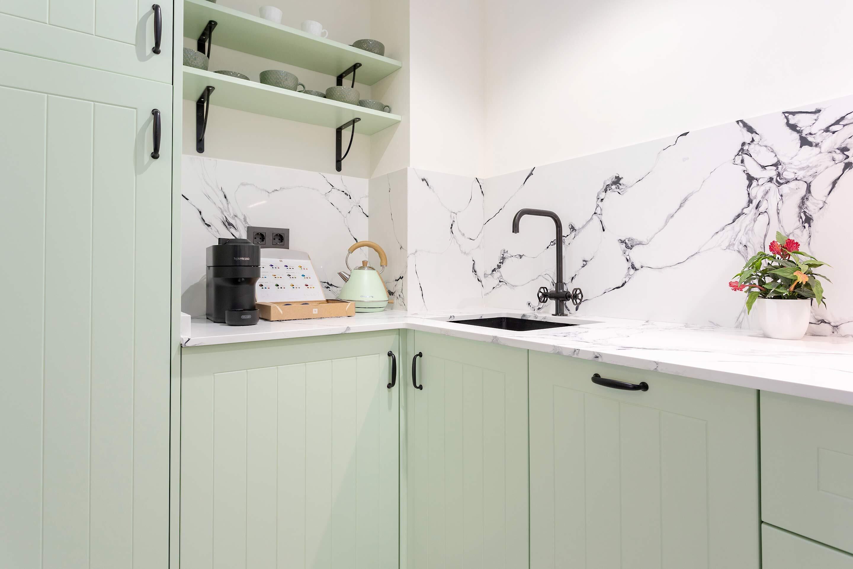 A kitchen with light green cabinets, a marble backsplash, a sink, a kettle, and a coffee maker.