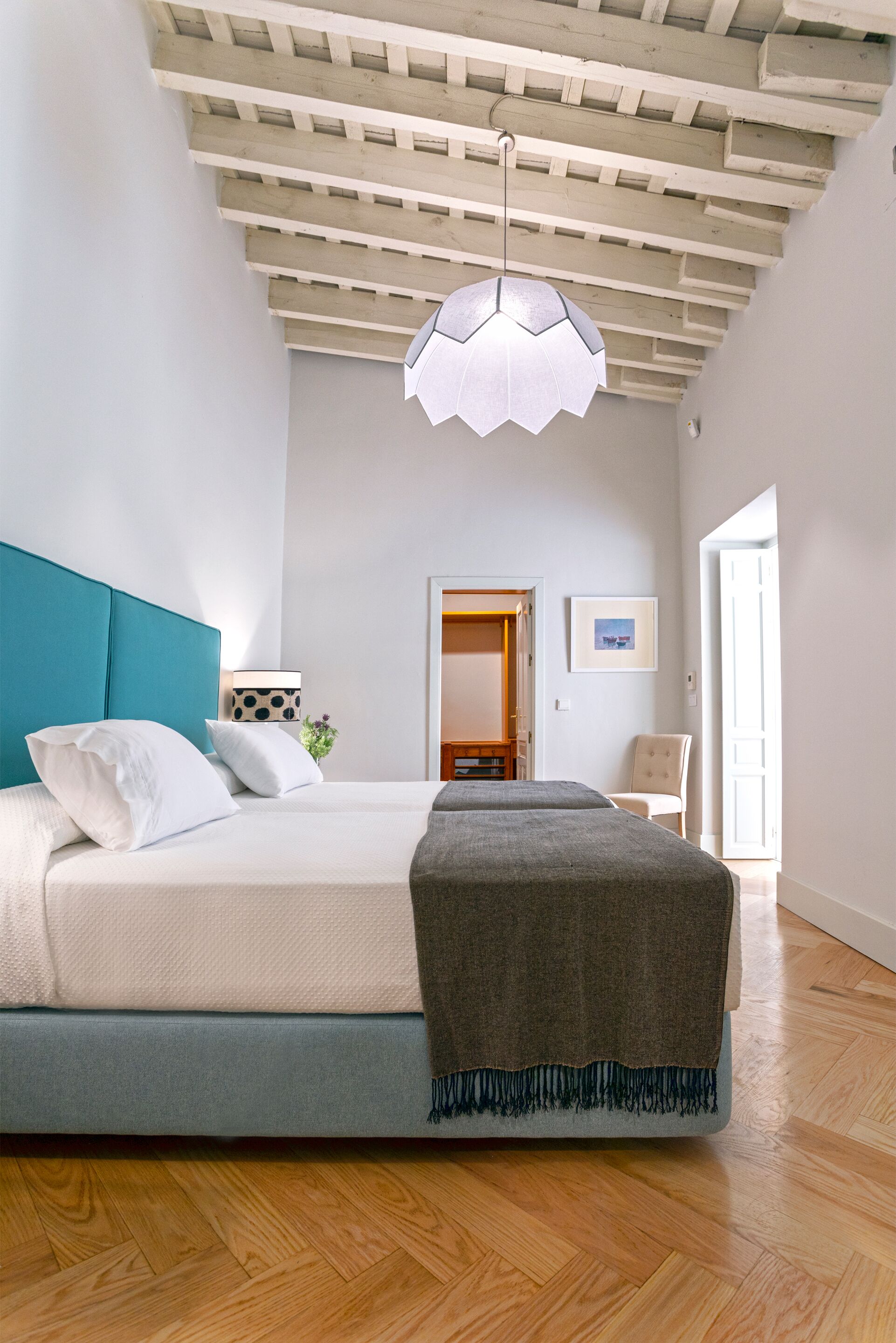 Ein Schlafzimmer mit einem Bett, einem blauen Kopfteil, einer Pendelleuchte, einer Holzbalkendecke, einer Tür und einem Sessel.