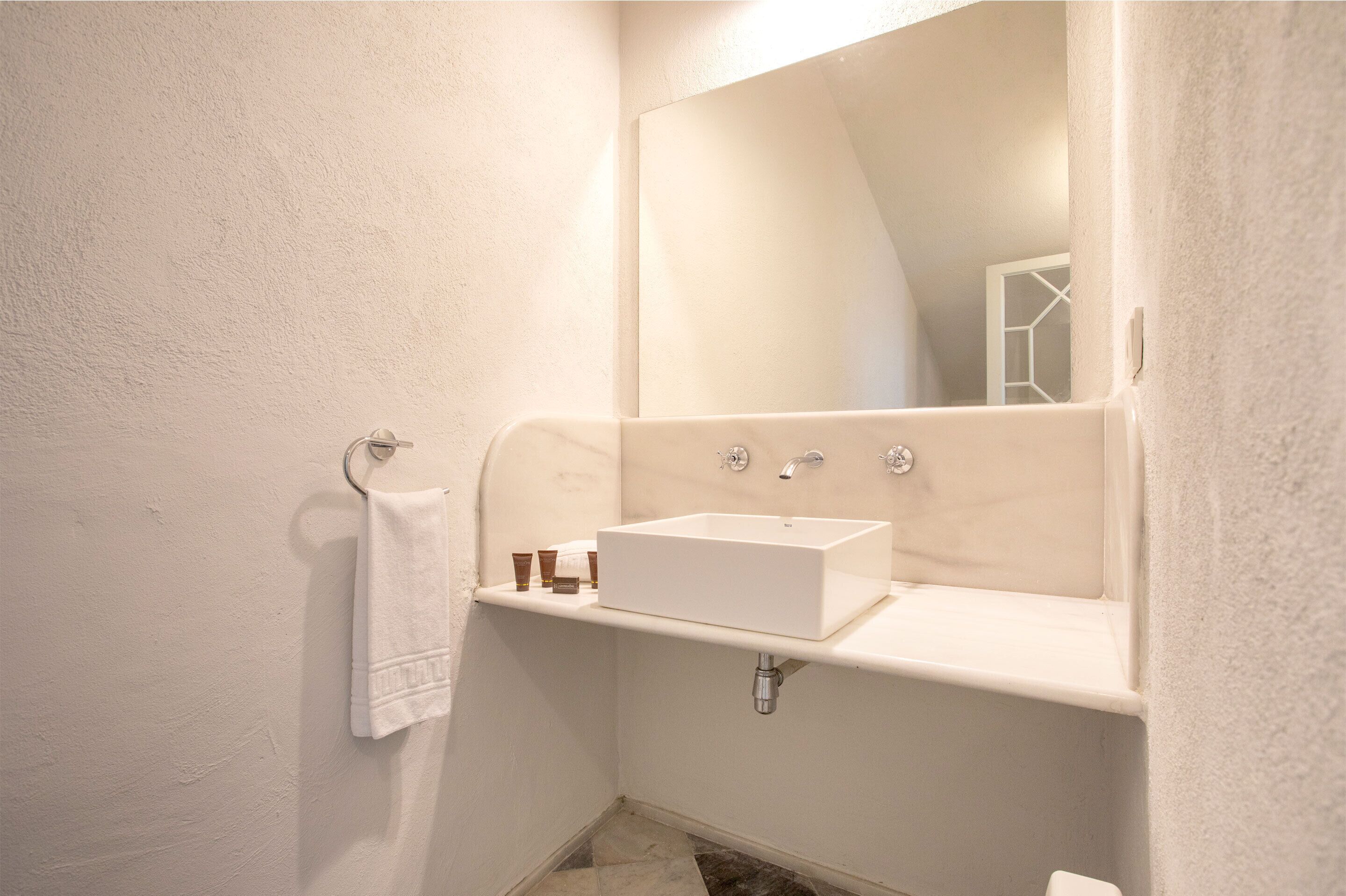 Un baño con lavamanos blanco, espejo y toallero.