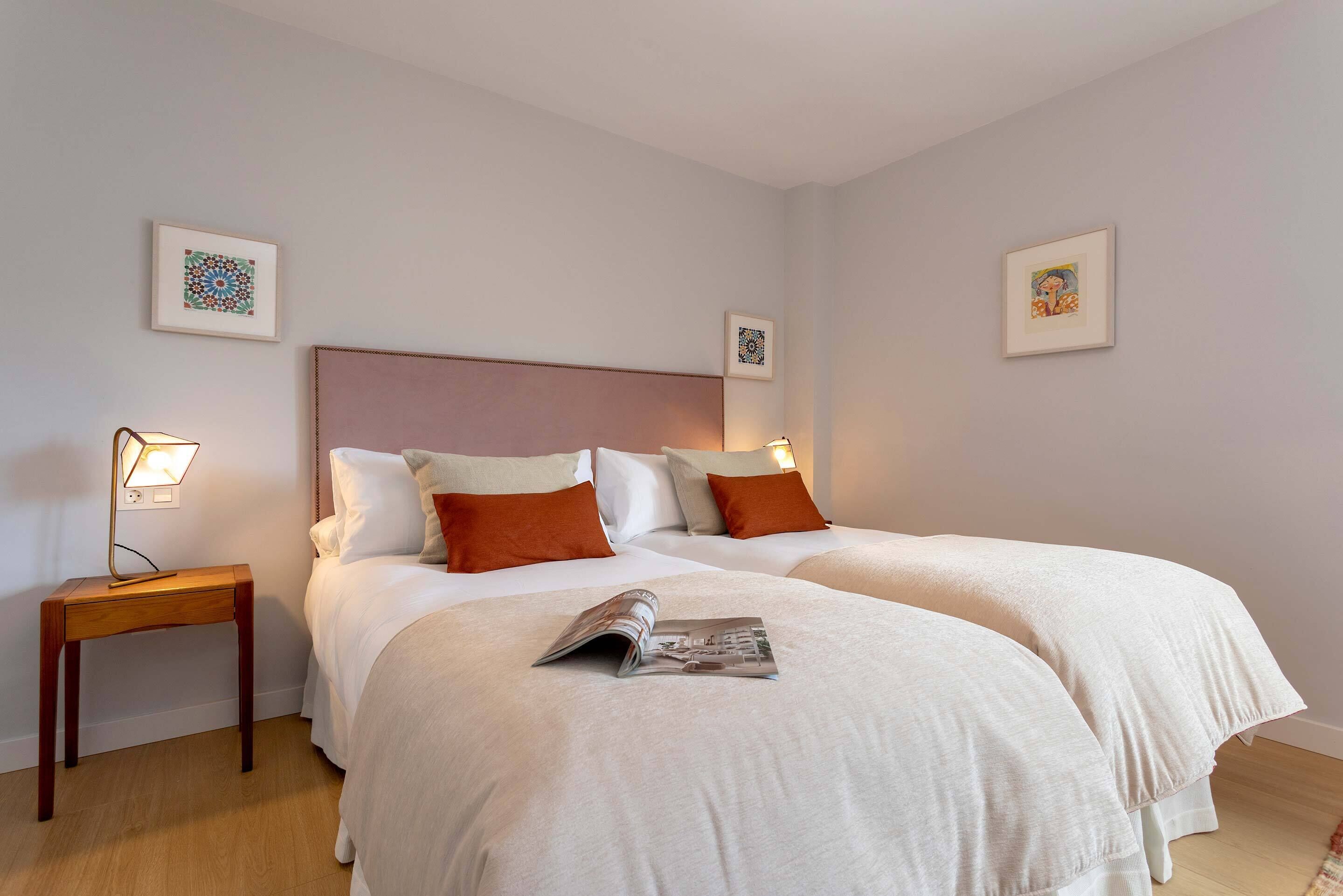 Un lit double avec une literie blanche et deux oreillers, une table de chevet avec une lampe, et deux œuvres d’art encadrées accrochées aux murs.