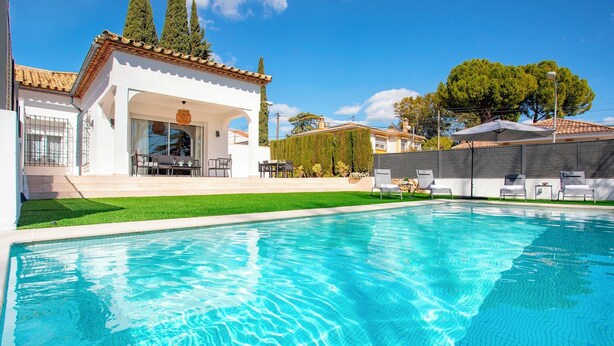A swimming pool with a clear blue surface, surrounded by a white building with a tiled roof and a covered outdoor seating area.