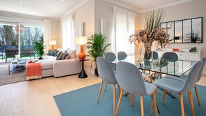 A modern living room with a sofa, a glass dining table, and chairs. There is a lamp, a potted plant, and a large window with curtains.