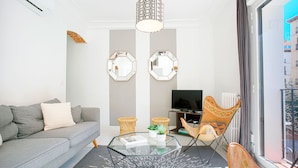Een moderne woonkamer met een grijze bank, een glazen salontafel en een televisie. Er hangen twee zeshoekige spiegels aan de muur, er staat een rieten fauteuil en er is een raam met gordijnen.