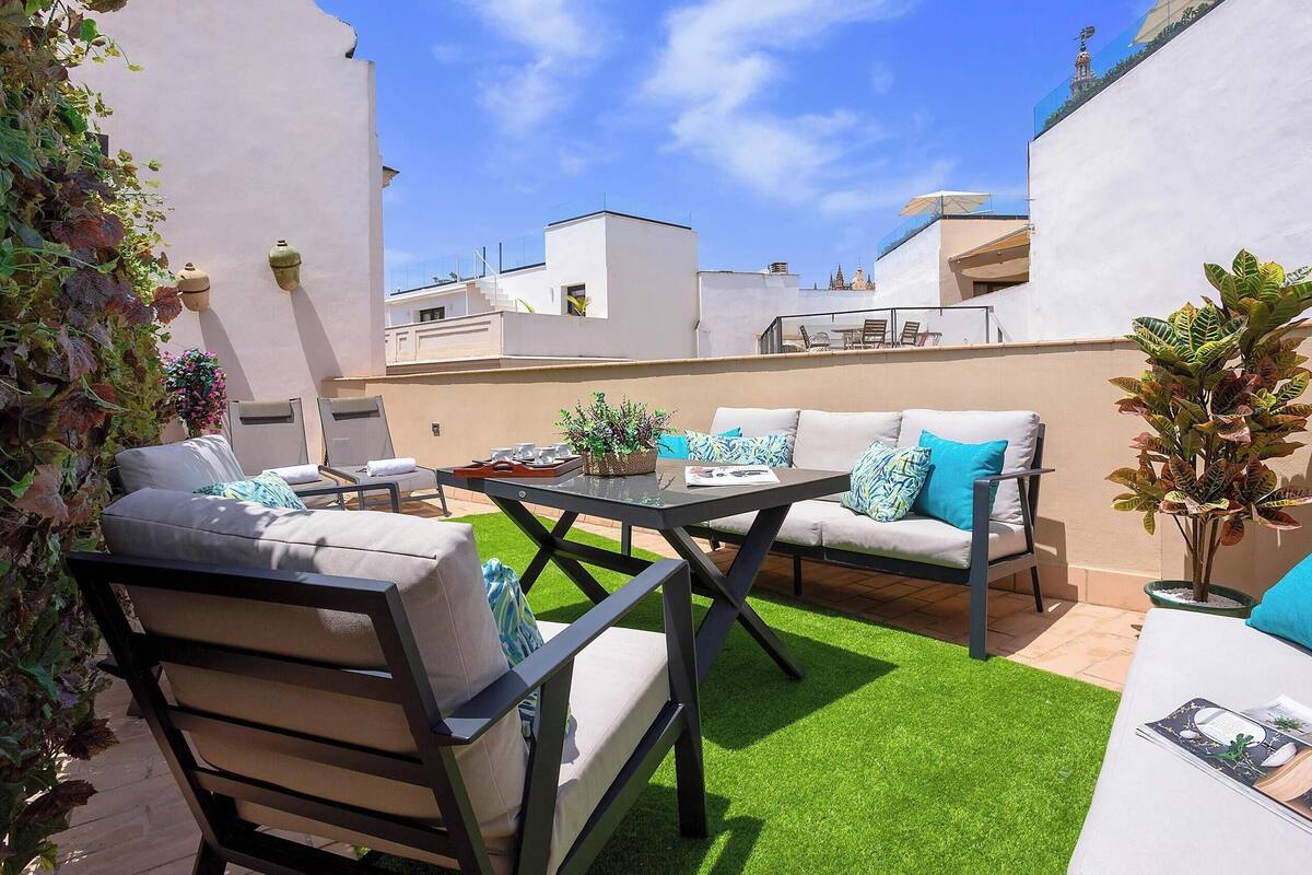 An outdoor patio with a sofa, chairs, and a table, surrounded by greenery and a clear sky.