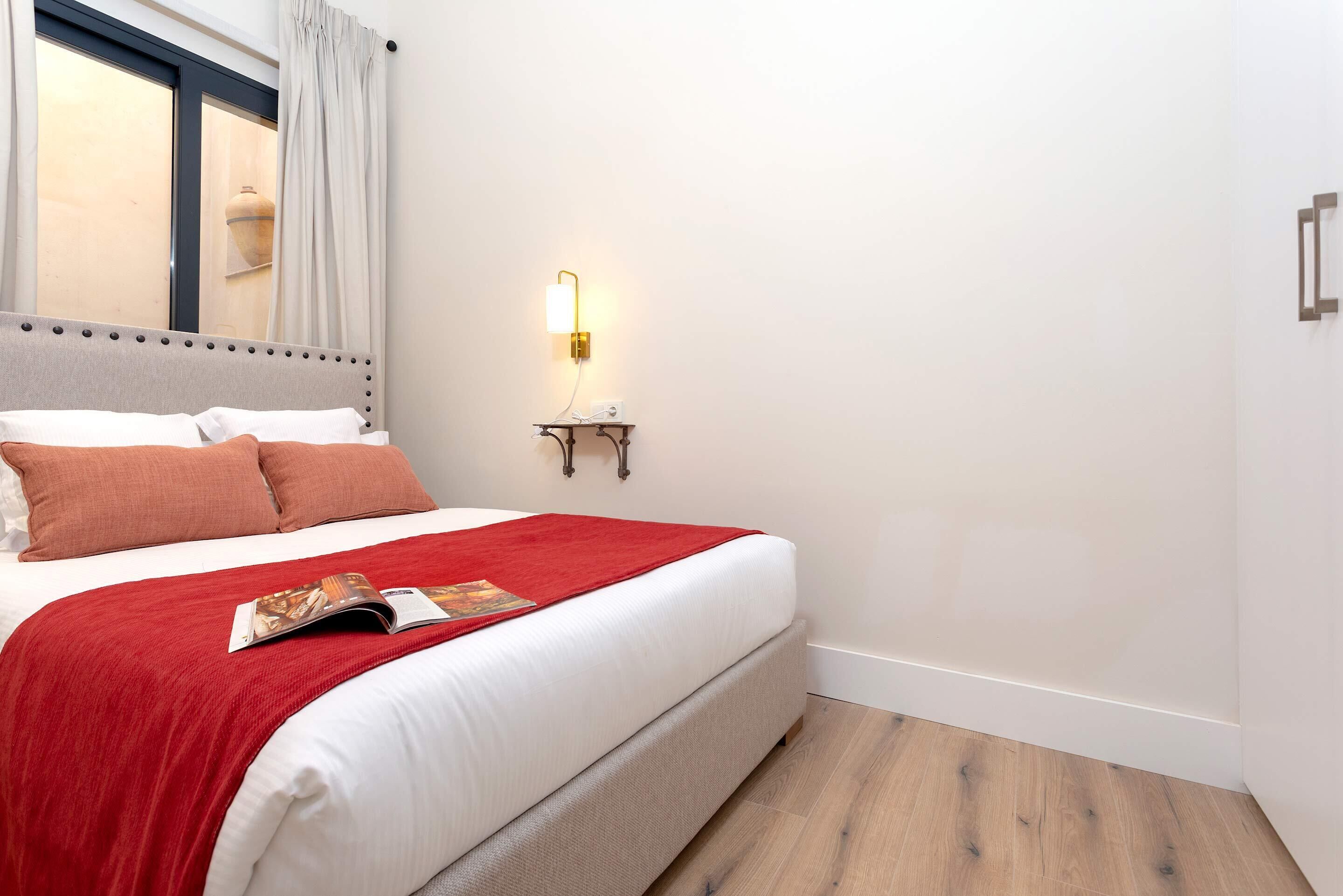 A neatly made bed with a red blanket and pillows, a bedside table with a lamp, and a window with curtains.