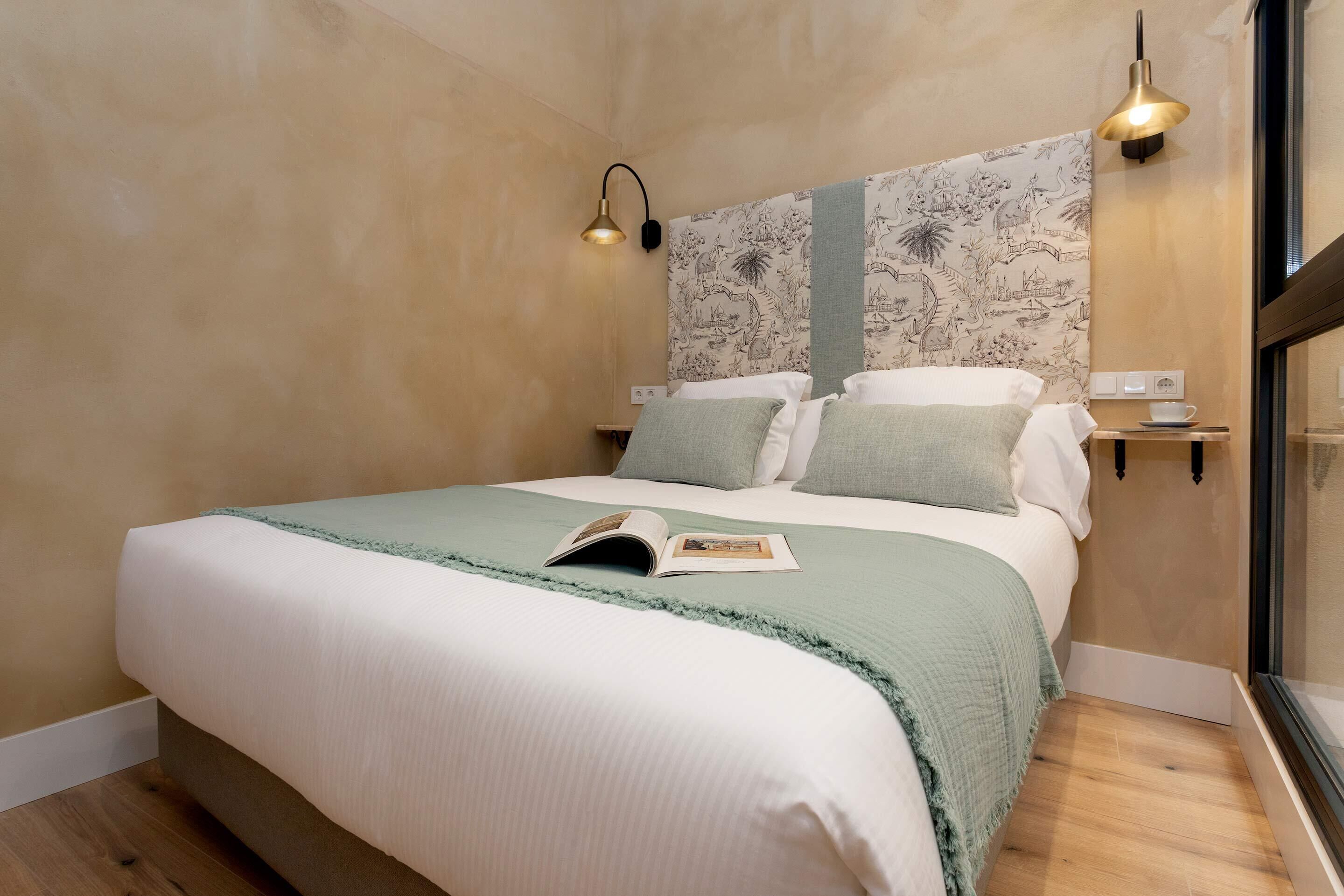 Un lit bien fait, recouvert d’une couverture verte, deux tables de chevet avec des lampes et un mur orné d’une tapisserie à motifs.