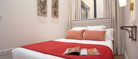 A neatly made bed with a red blanket and pillows, a magazine on the bed, and two wall-mounted paintings.