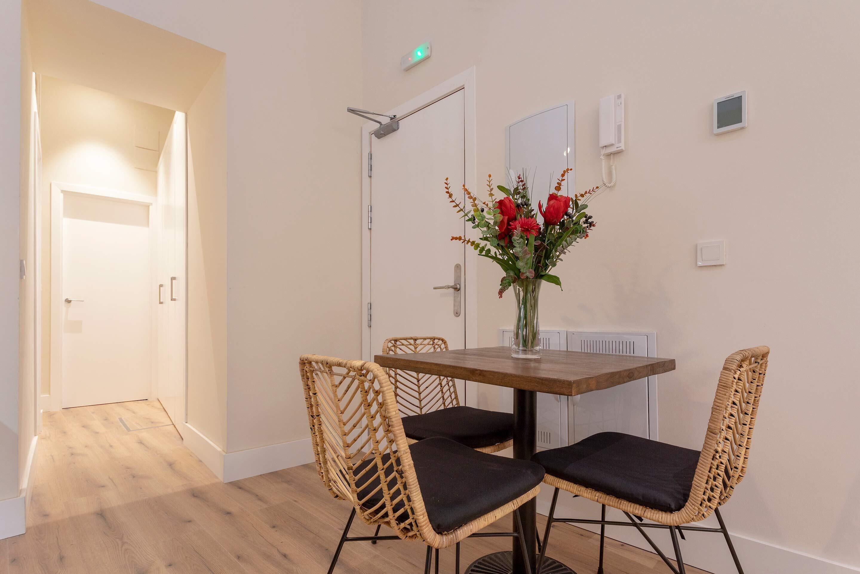 A dining area with a wooden table and wicker chairs, a vase with flowers, and a door with a light switch.