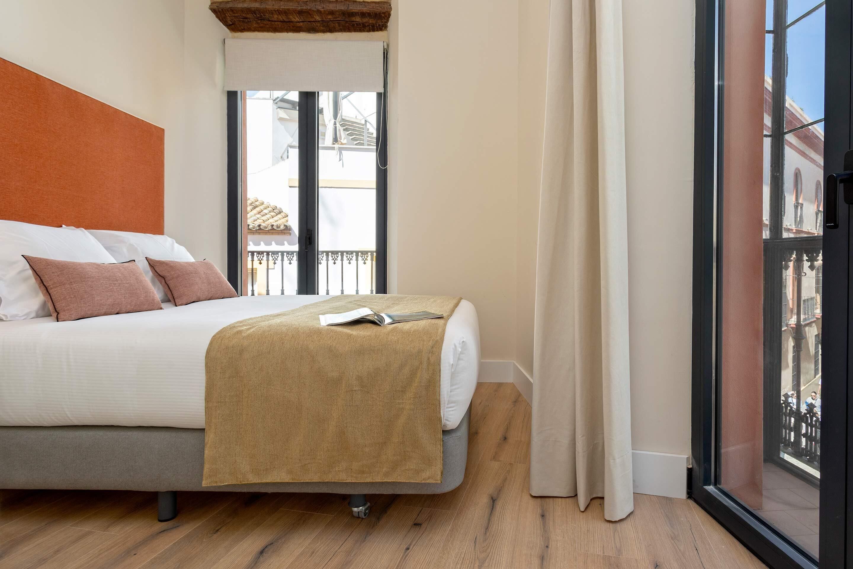 A bedroom with a bed, a window with a view of buildings, and a balcony.
