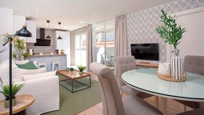 Ein modernes Wohnzimmer mit Glastisch, bequemen Sitzgelegenheiten, einer Küche im Hintergrund und einem an der Wand montierten Fernseher.
