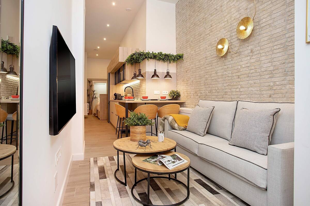 A modern living room with a sofa, coffee table, and TV. There is a kitchen in the background with a brick wall and pendant lights.