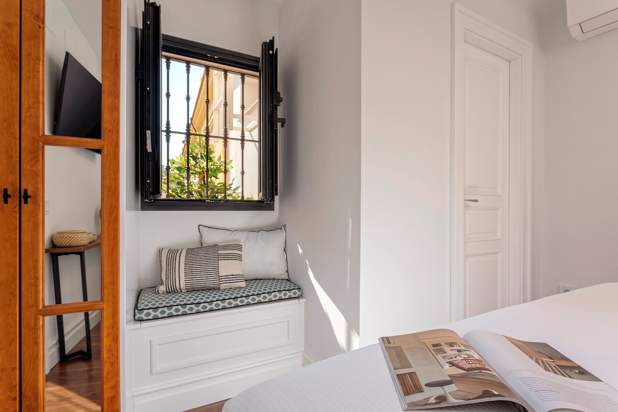 Una habitación con una cama, una ventana con contraventanas negras, un banco con cojines y una revista sobre la cama.