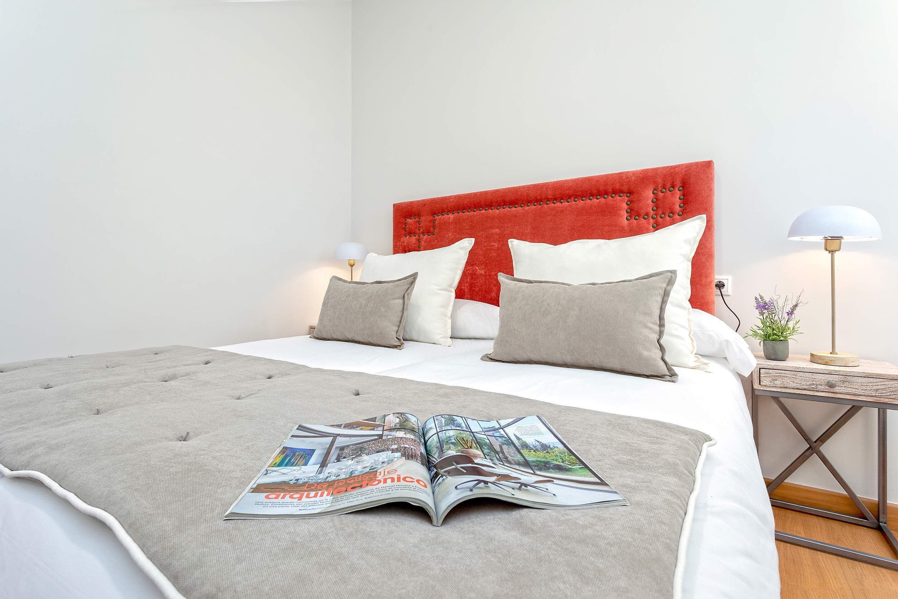 Una cama bien hecha con cabecera roja, almohadas grises y blancas, y una revista sobre la cama.
