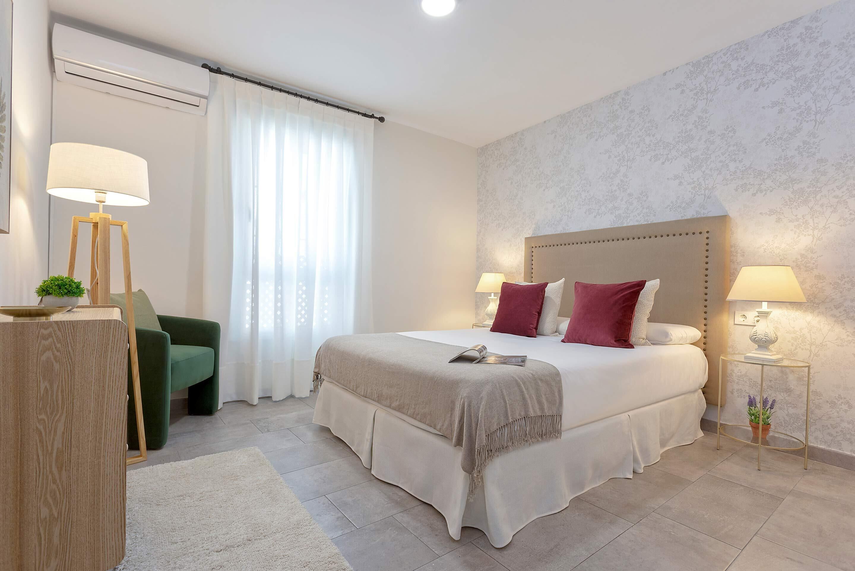 Un dormitorio ordenado con una cama grande, lámparas de noche, un sillón verde y una mesita con una planta.