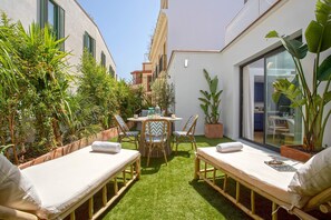 Un pequeño patio al aire libre con dos sillones, una mesa y sillas, rodeado de abundante vegetación y vistas al interior.