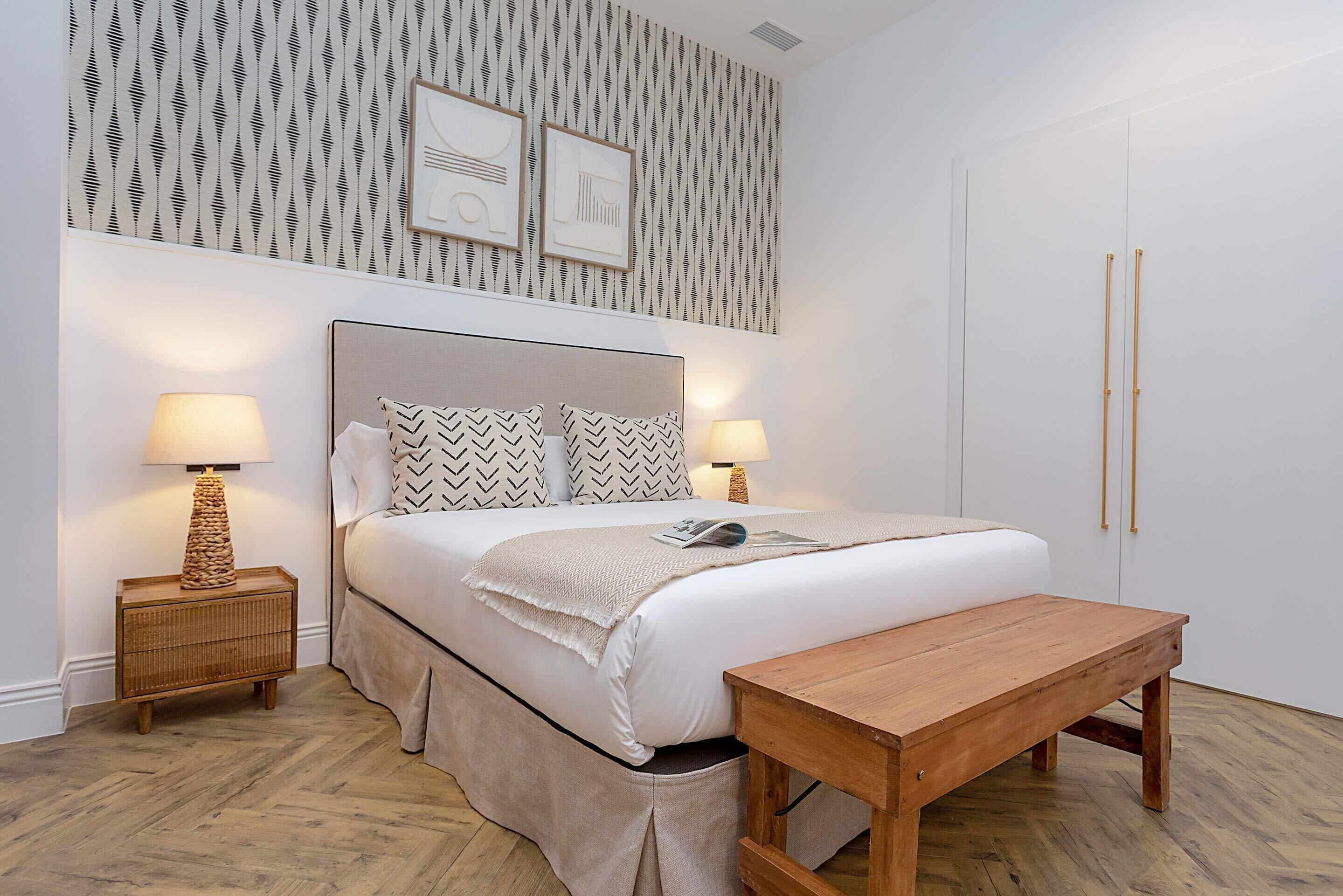 Quarto bem organizado com cama, duas mesas de cabeceira com abajures, um banco de madeira e papel de parede estampado.