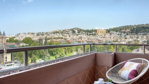 Ένα μπαλκόνι με μια καρέκλα από μπαμπού, ένα μικρό τραπεζάκι με ένα περιοδικό και θέα στην πόλη, με τα κτίρια και την πρασινάδα της.