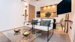 A modern living room with a glass coffee table, white sofas, and a kitchen area with wooden cabinets and a flat-screen TV. - Genteel Home Teodosio (Seville)