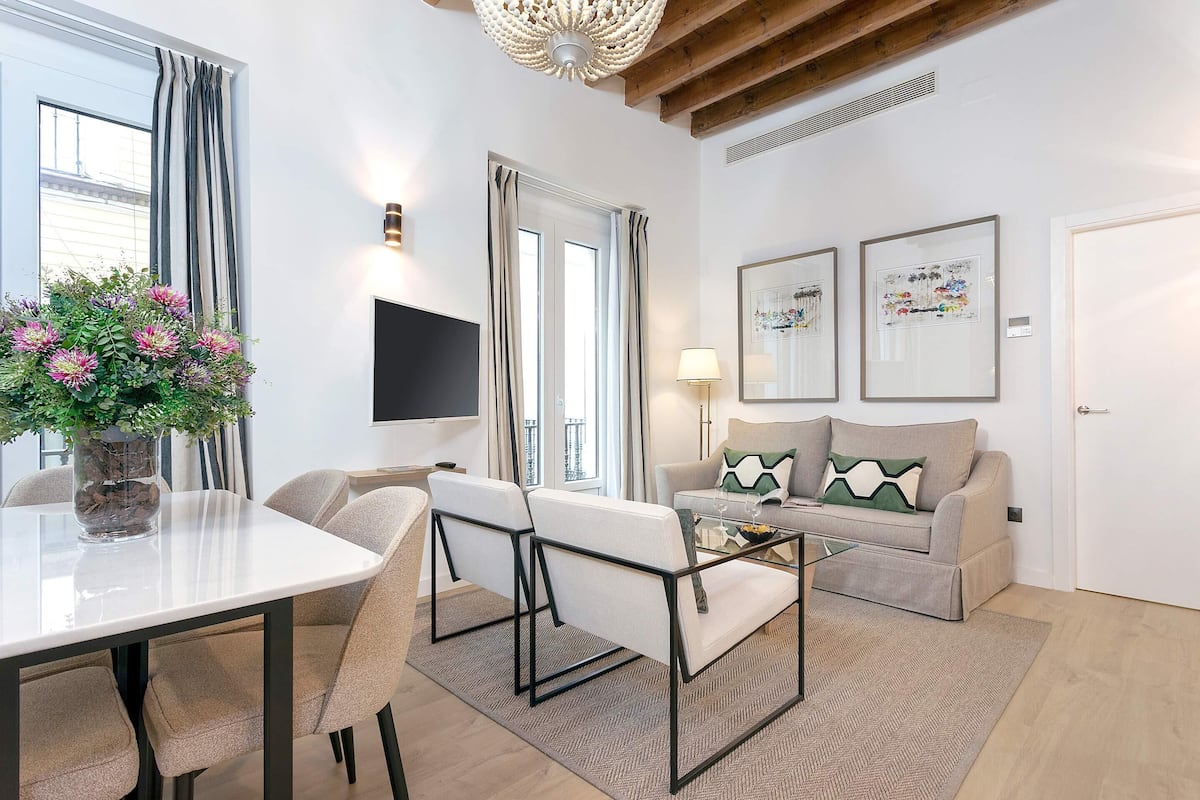 Una sala de estar moderna con una mesa de comedor, sillas, un sofá y una mesa de centro. Hay dos obras de arte enmarcadas en la pared, un televisor de pantalla plana montado sobre el sofá y una planta en maceta sobre la mesa.