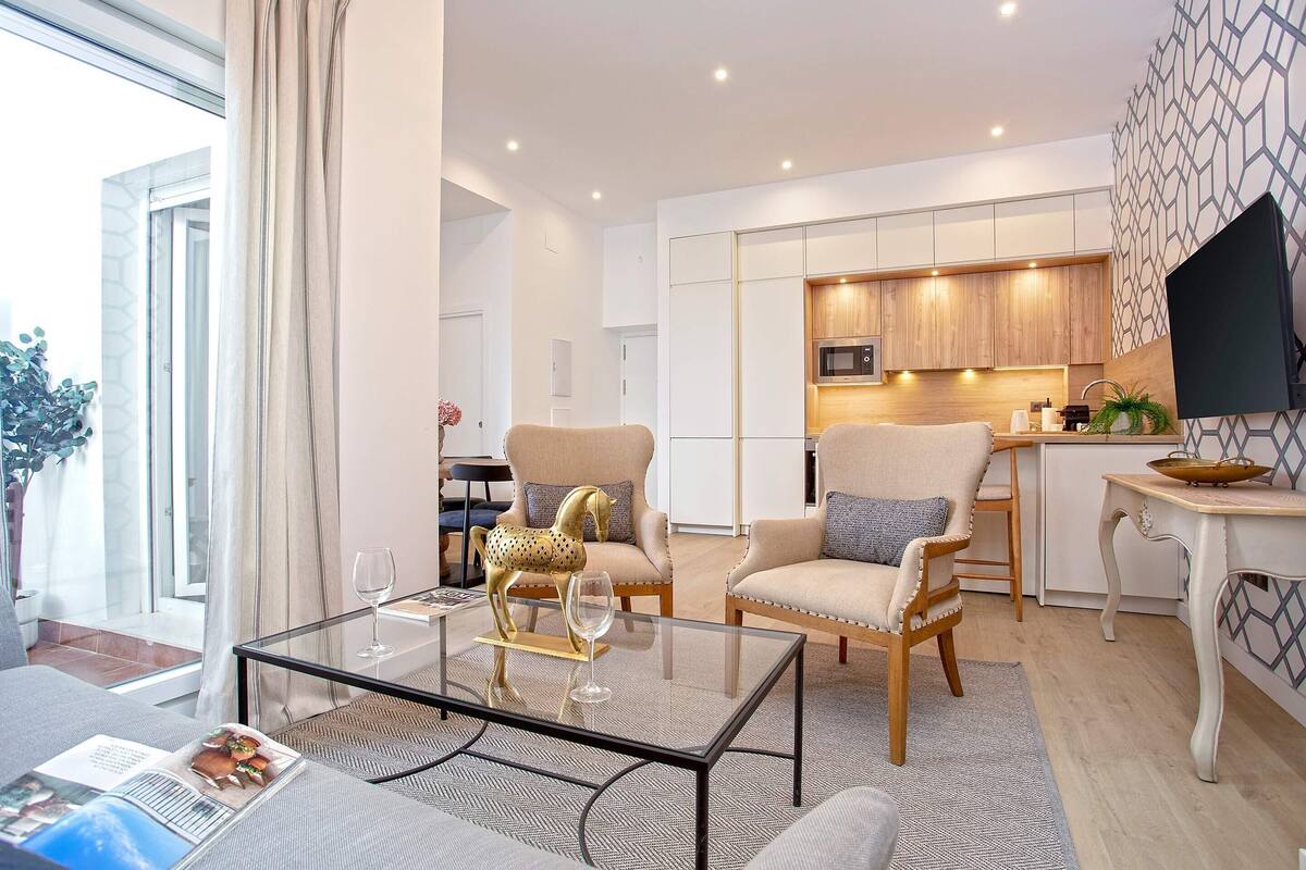 Una sala de estar moderna con una mesa de centro de vidrio, dos sillones, un sofá y vistas al área de la cocina.
