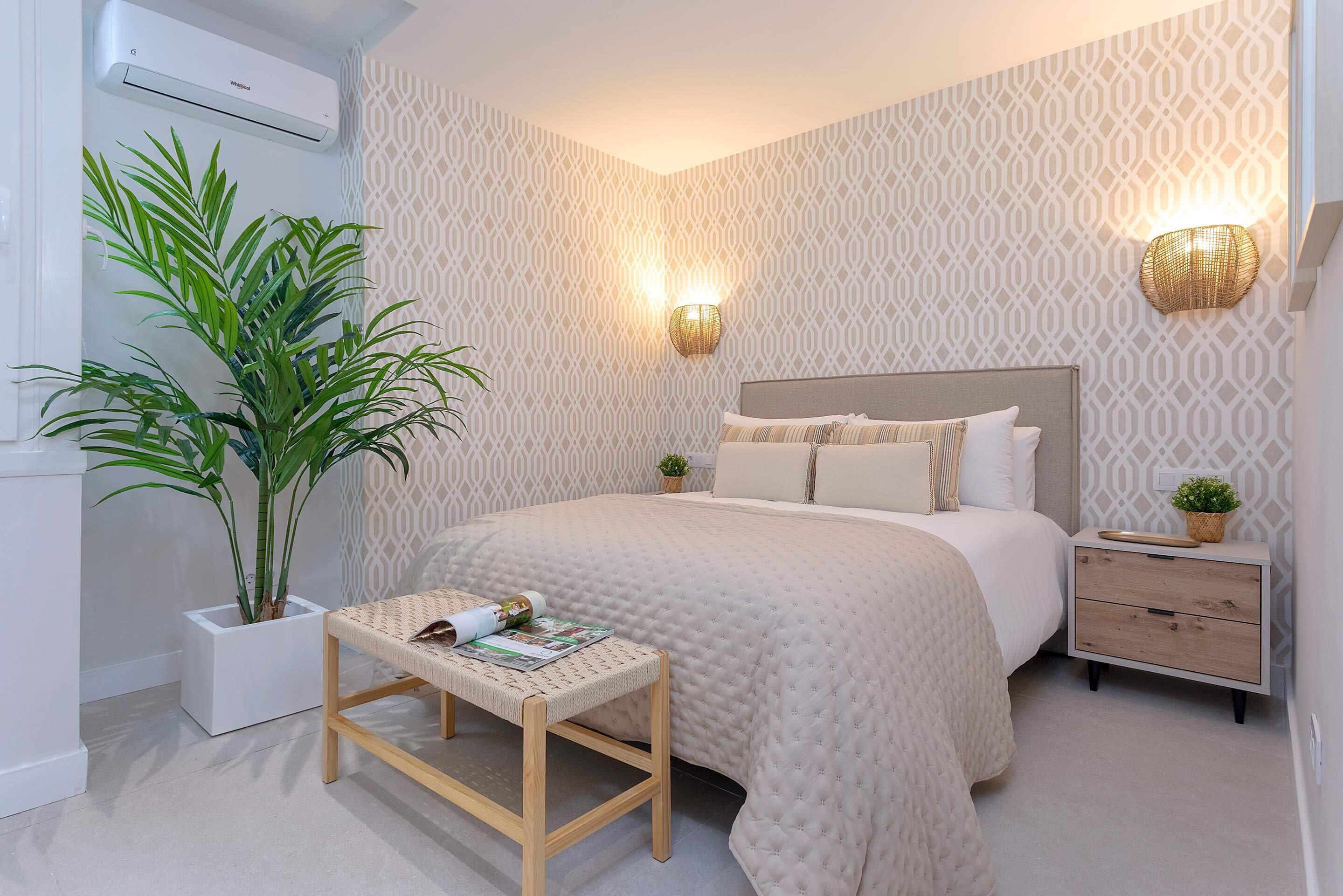 A bedroom with a bed, bedside tables, a wall-mounted air conditioner, a potted plant, and a wall-mounted light fixture.