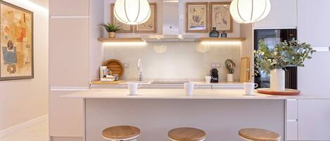 Una cocina moderna con una isla central, taburetes de bar, iluminación colgante y obras enmarcadas en las paredes.