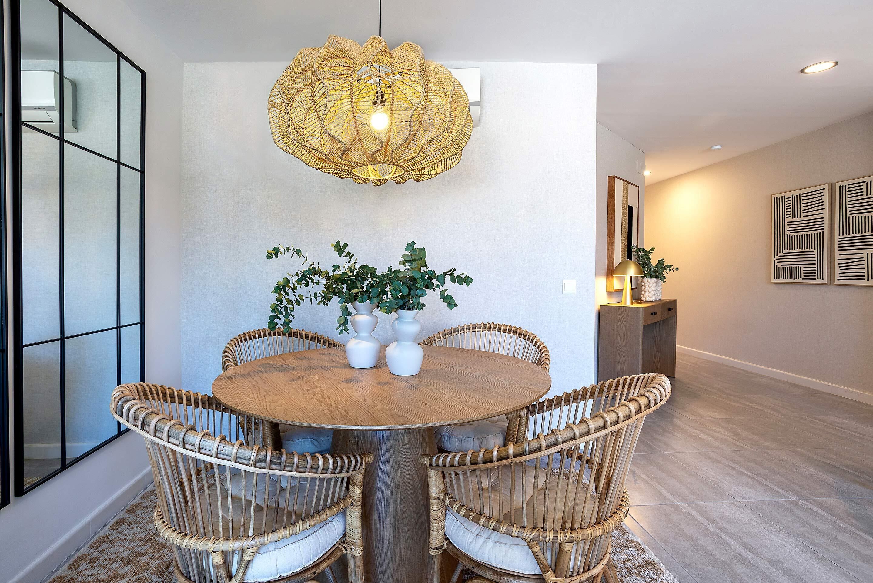 Un comedor con una mesa redonda de madera rodeada de sillas de mimbre, un lámpara colgante y plantas en macetas.