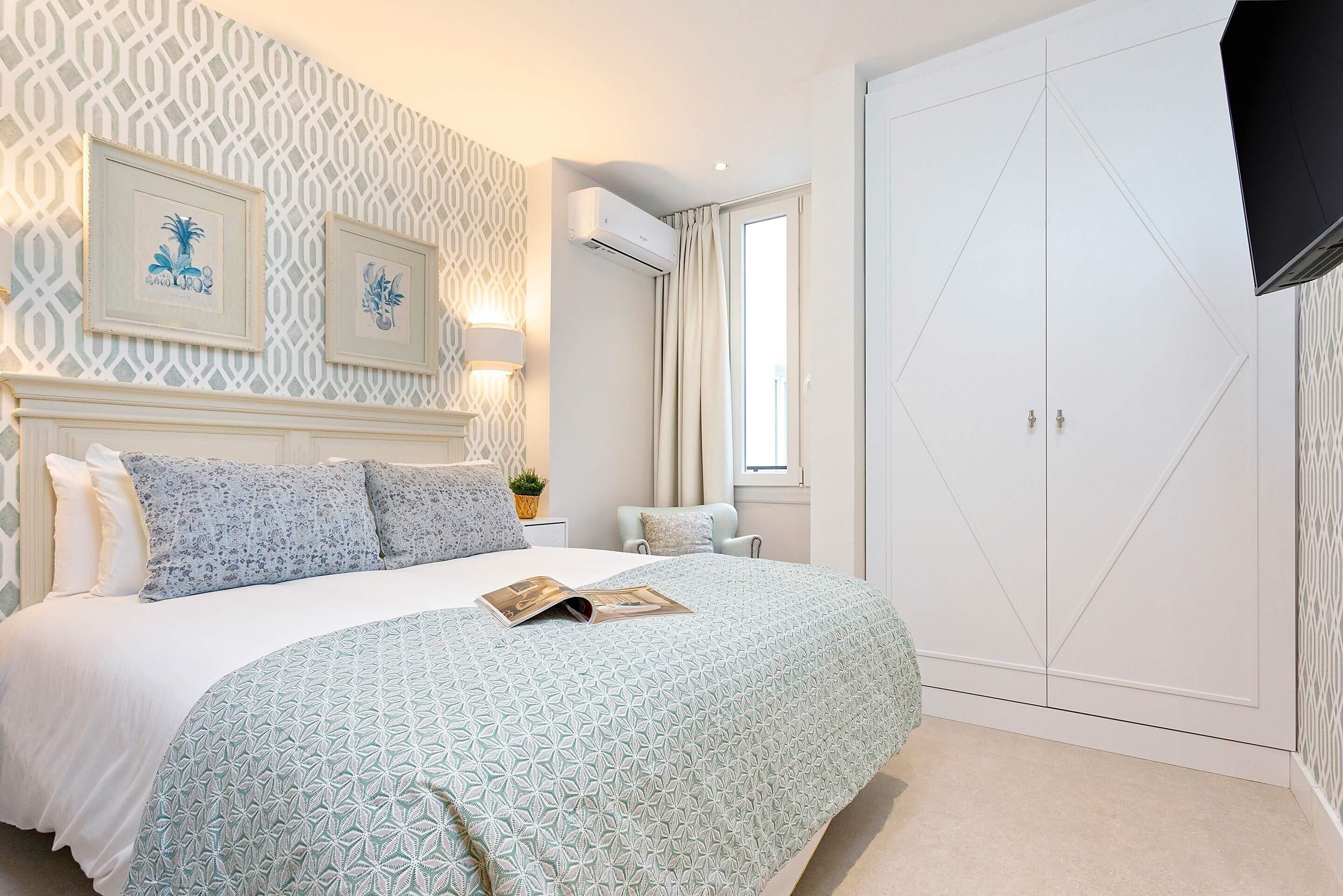 Un dormitorio con cama, armario, mesita de noche, aire acondicionado de pared y cuadros enmarcados.