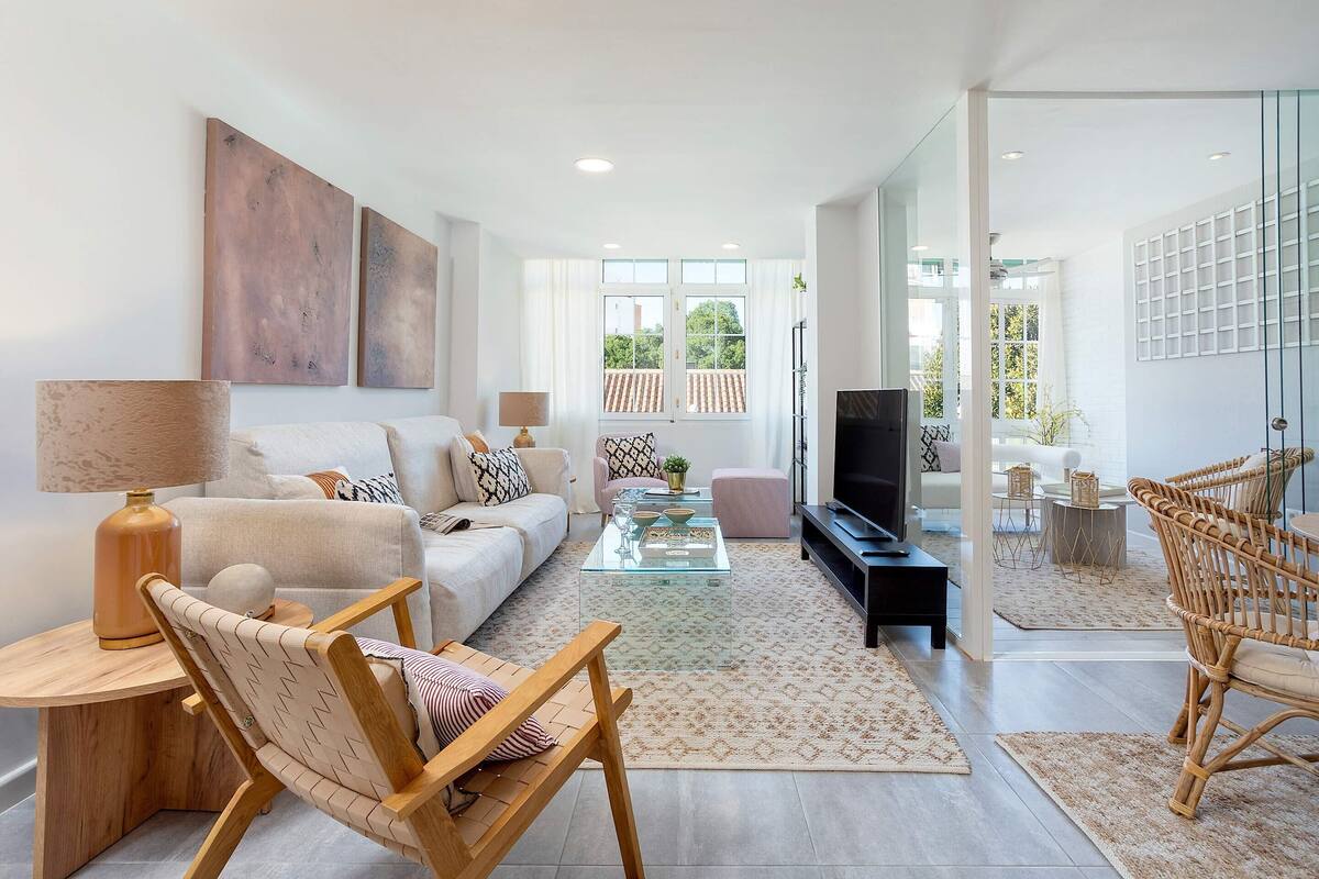 A modern living room with a sofa, coffee table, and chairs. There is a television and a window with a view of greenery.