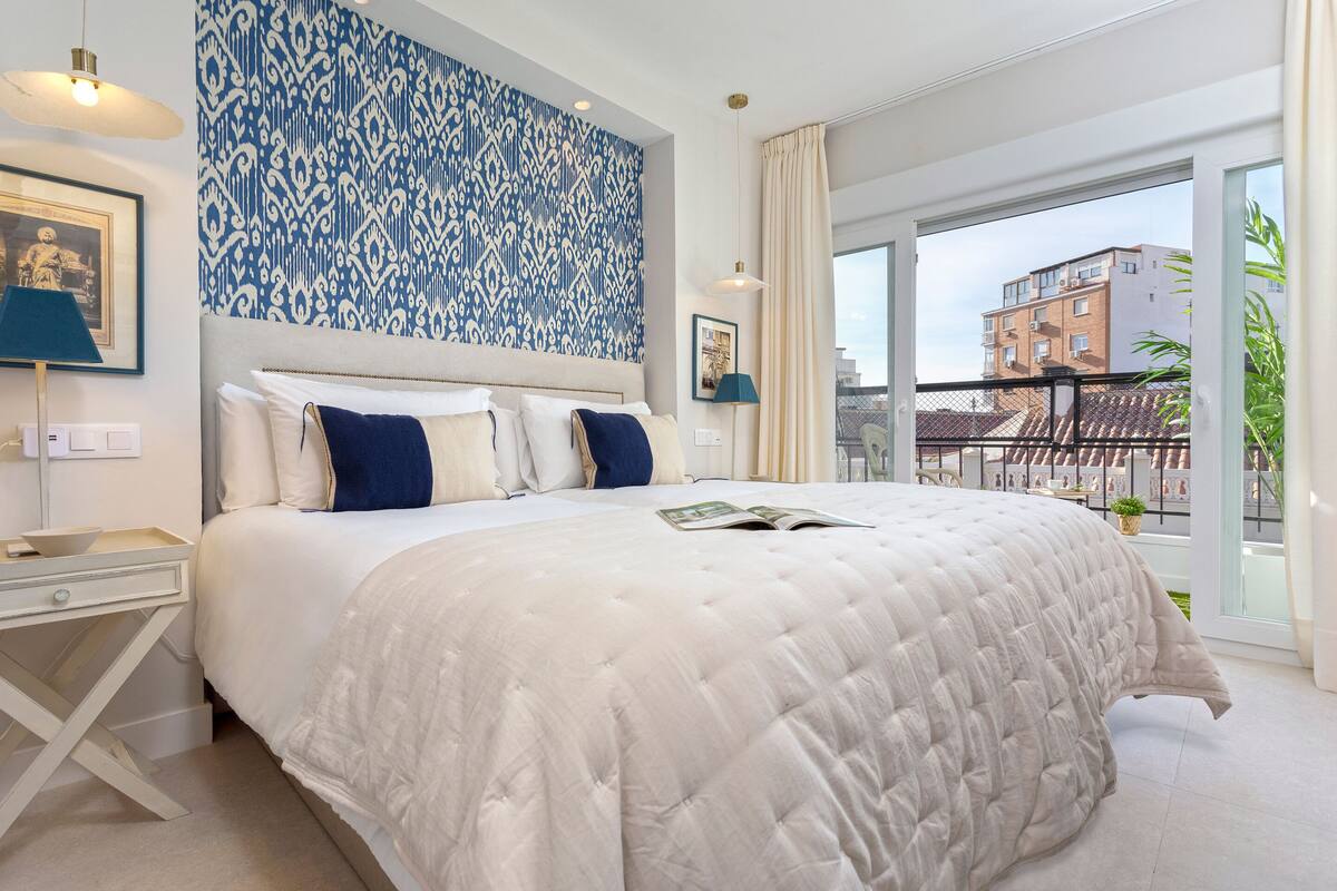 A modern bedroom with a large bed, a patterned wall, and a view of buildings through a large window.