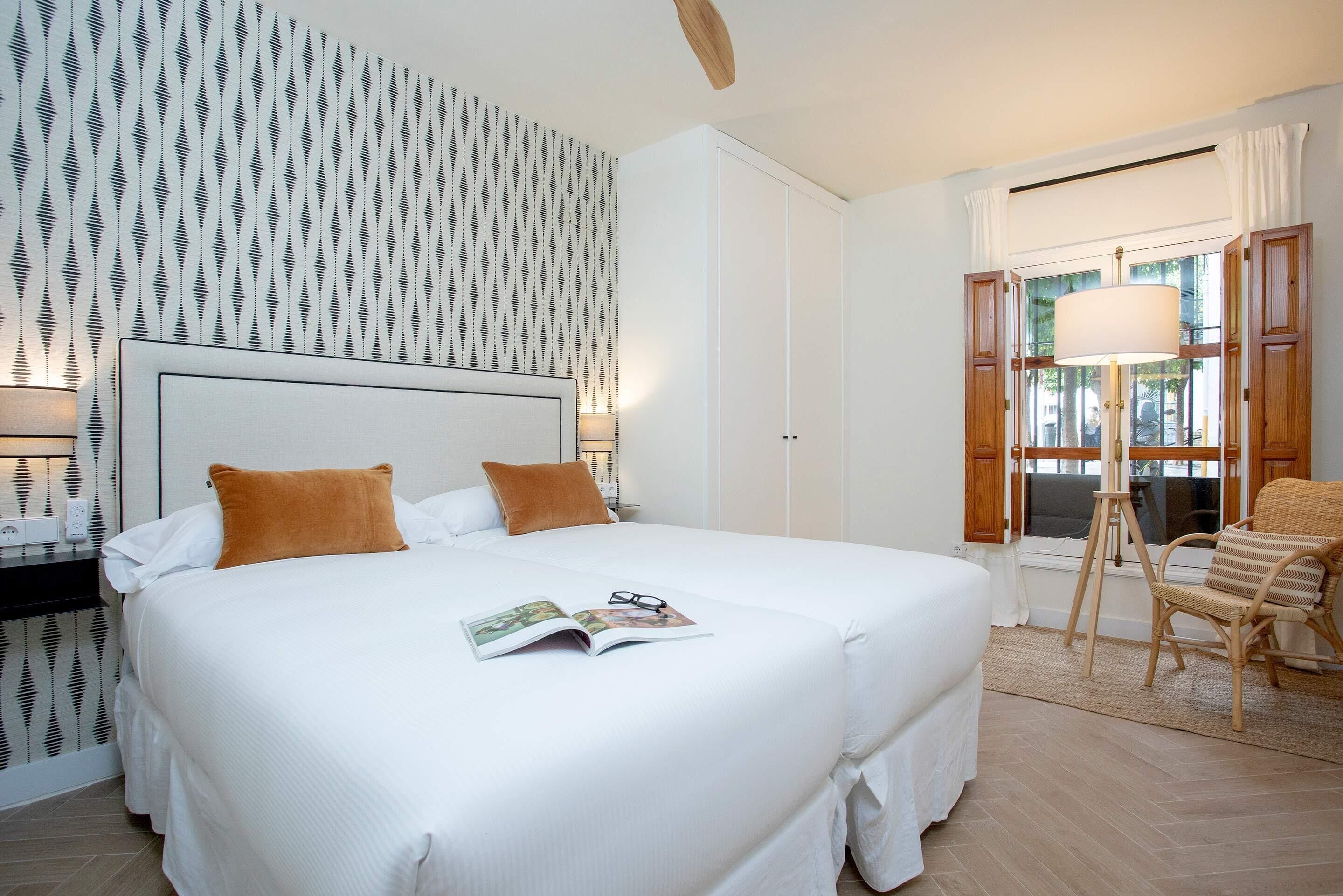 Una cama bien hecha con una revista y gafas, una mesita de noche con una lámpara, y una ventana con cortinas.